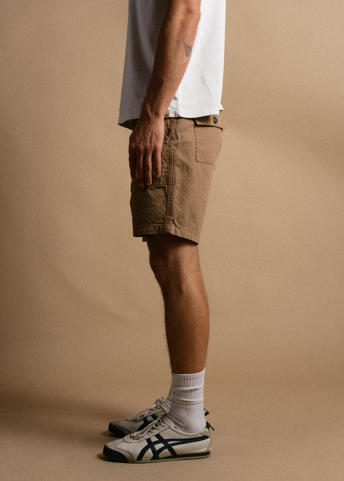Man in white shirt, beige shorts, and sneakers stands sideways against a plain background, showcasing casual summer style.
