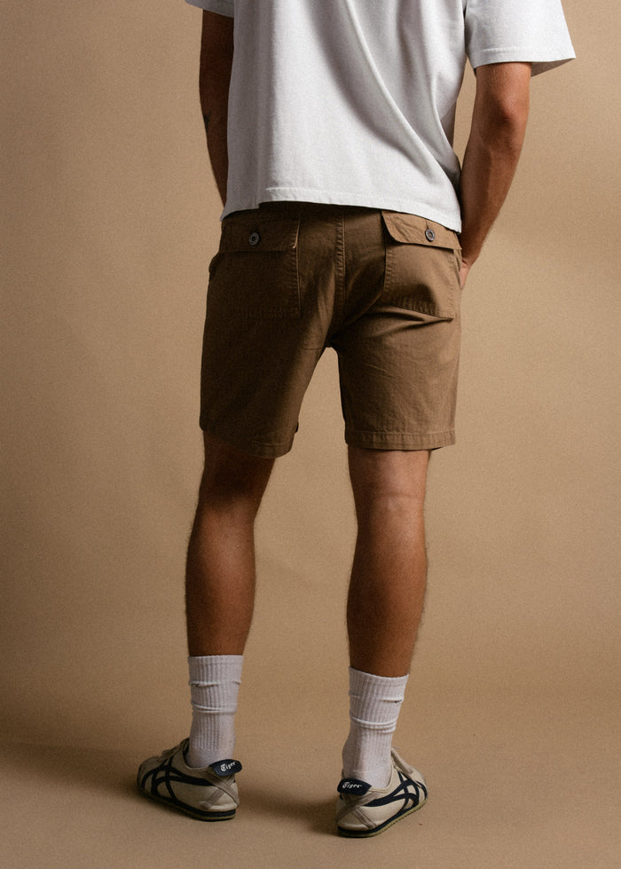 Man in white shirt and brown shorts, facing away, stands on beige background. Wearing white socks and sneakers. Casual style.