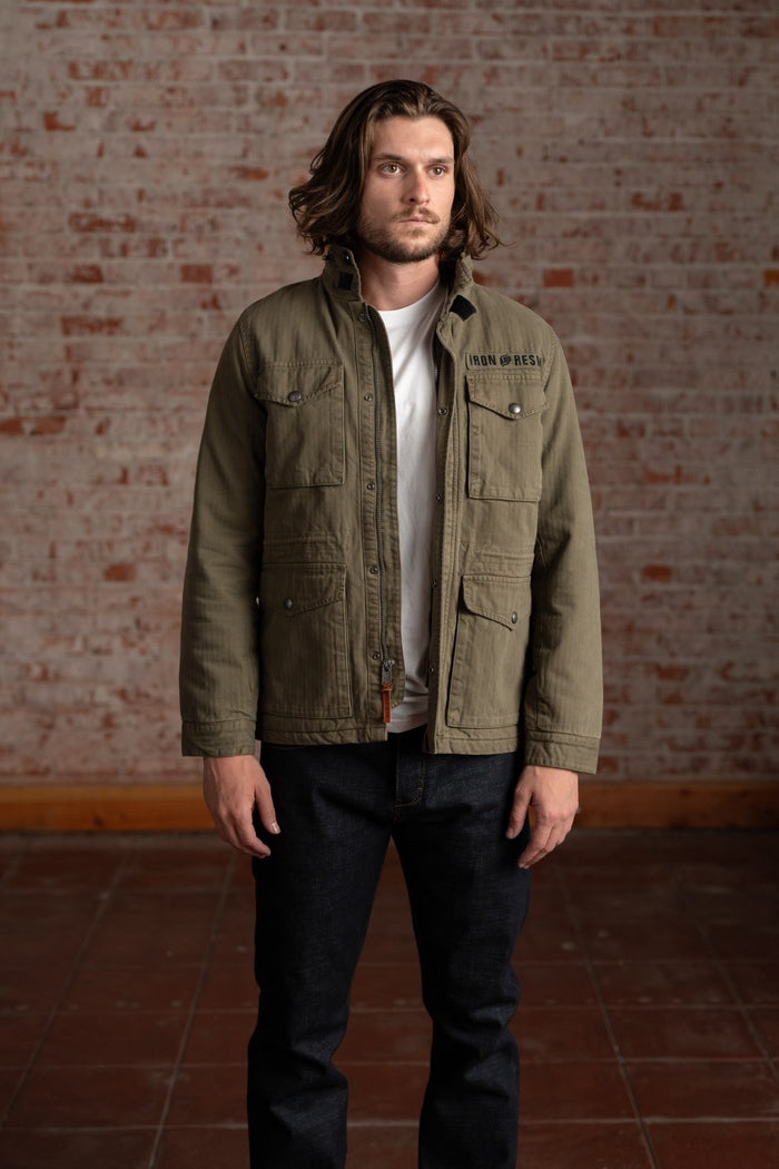 Man in olive jacket and jeans stands against a brick wall, wearing a white shirt. The jacket has "Iron Resin" text on it.