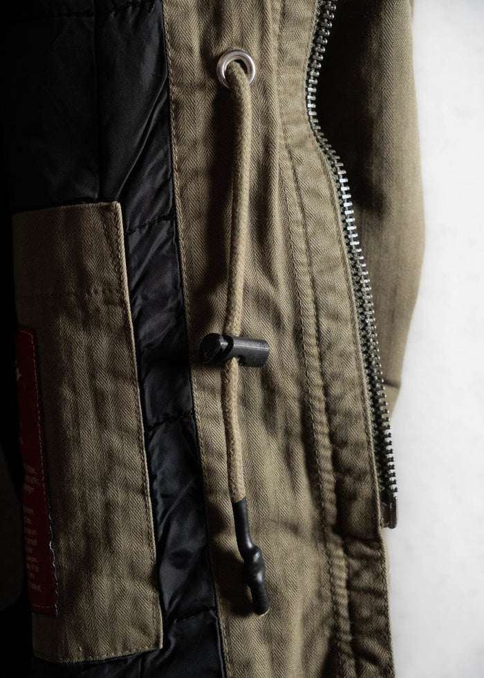 Close-up of a khaki jacket with a black drawstring, metal eyelet, and silver zipper, highlighting its detailed stitching.