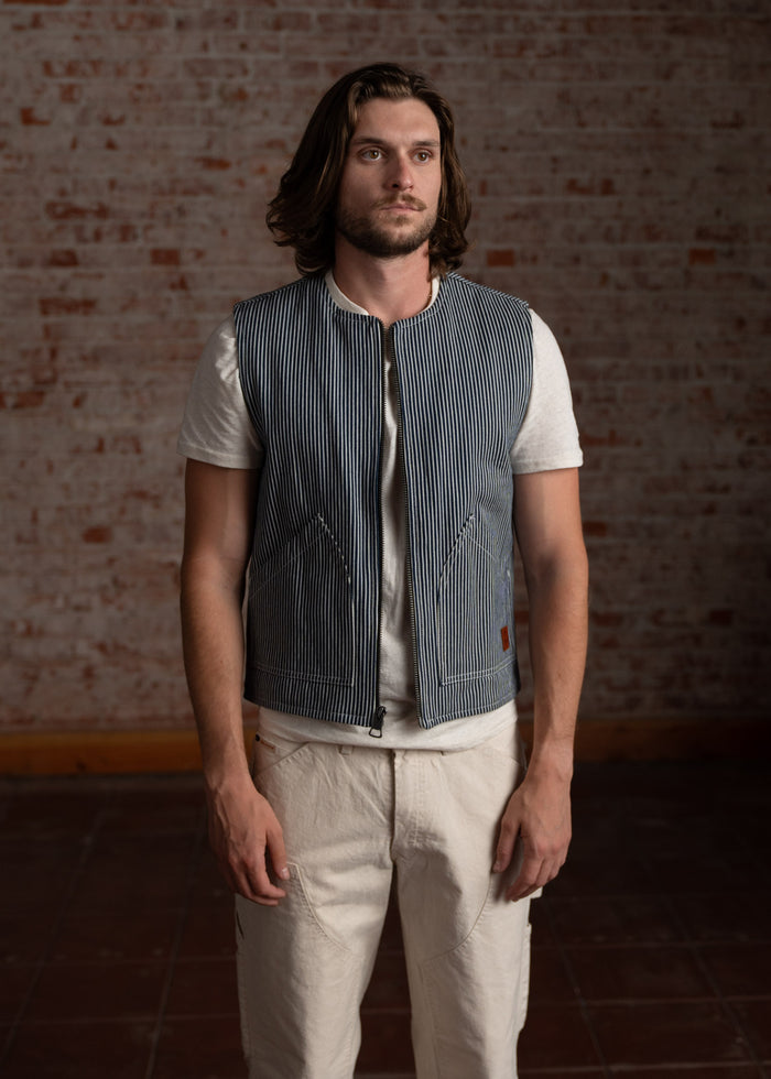 Man in a striped vest and white shirt stands against a brick wall, looking forward. Casual and relaxed style.