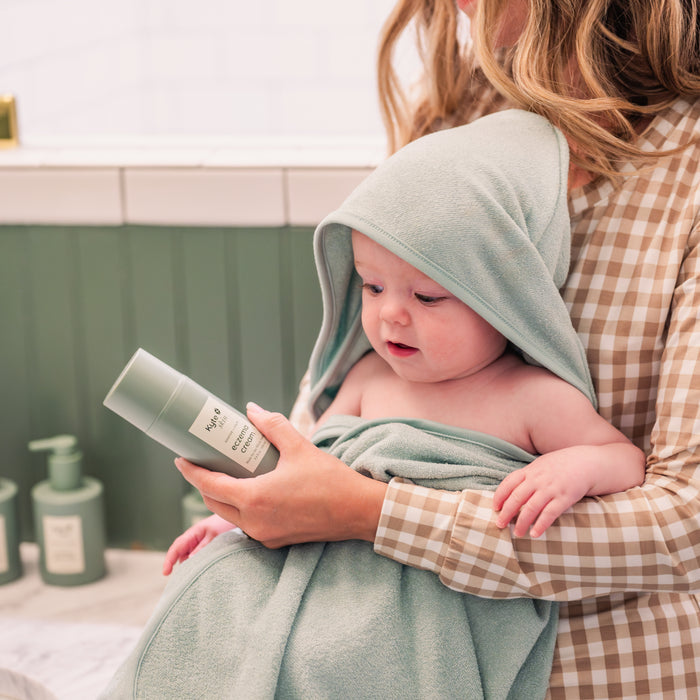 Mom holding baby wrapped in a sage green hooded bamboo terry towel holding a bottle of kyte skin eczema cream