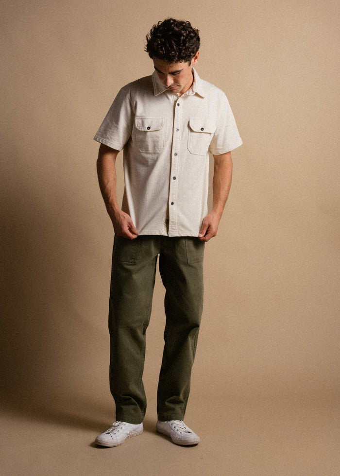 Man in a beige shirt and green pants stands on a tan background, looking down, wearing white sneakers.