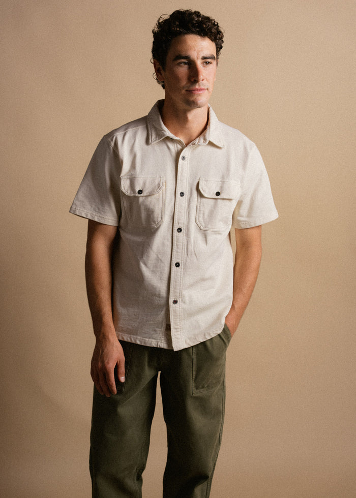 Man in beige shirt and green pants stands against a neutral background, looking to the side with a calm expression.