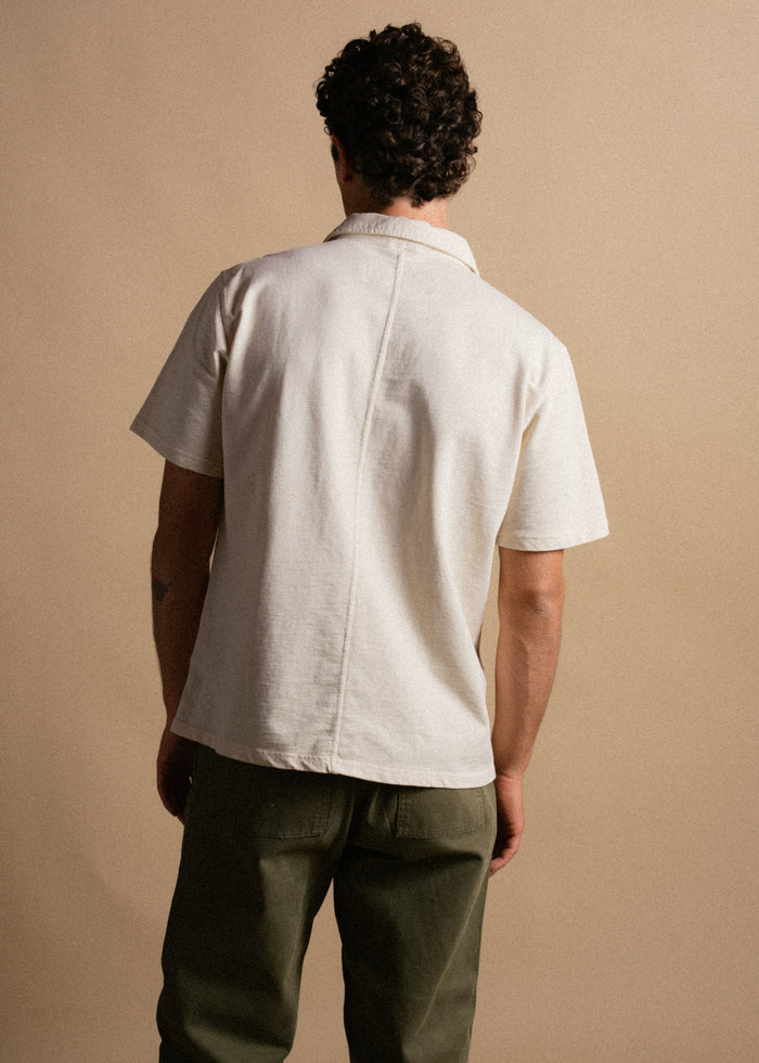 Man in a white short-sleeve shirt and green pants stands with back to camera against a beige background, showcasing casual style.