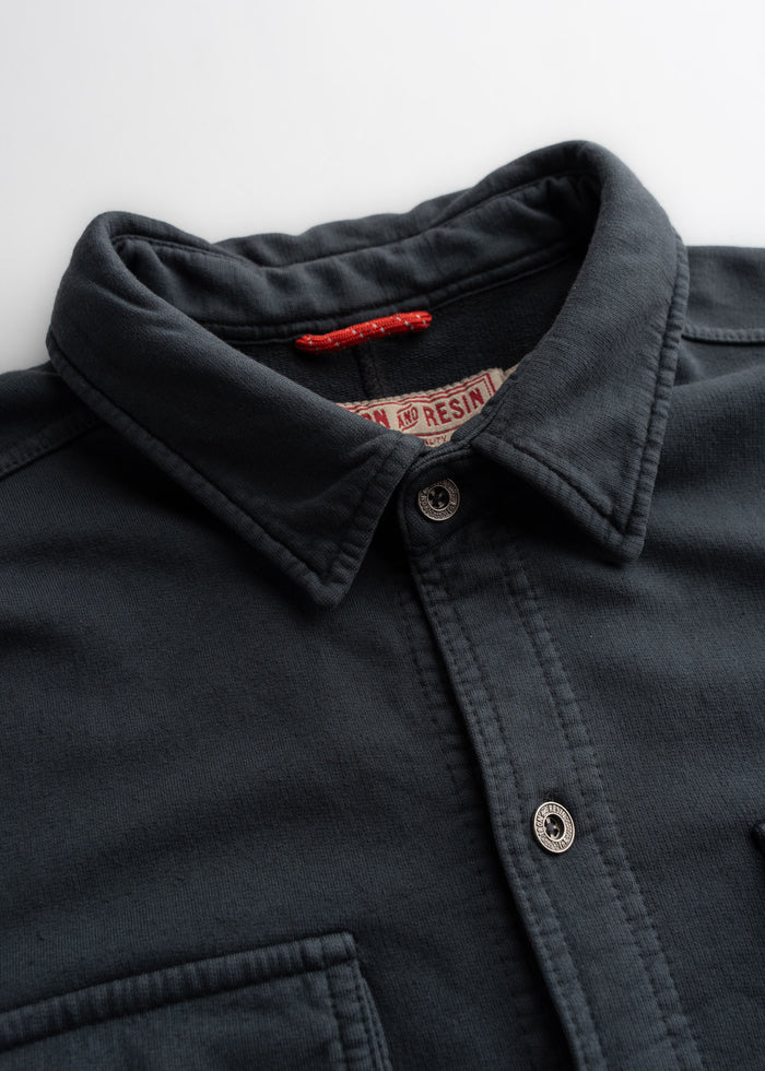 Dark button-up shirt with a collar, featuring a red tag inside. The fabric appears soft and textured, with visible stitching.