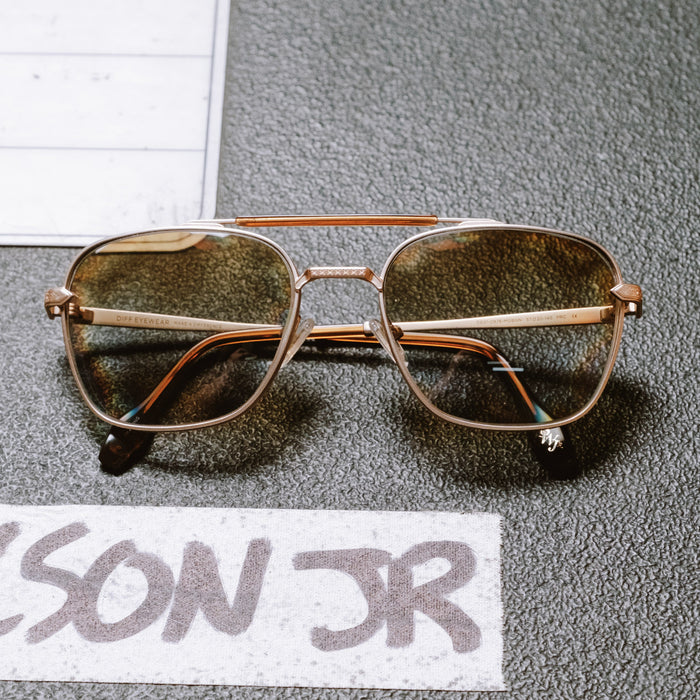 still life - diff eyewear x stephen wilson jr. i'm a song navigator sunglasses with a gold metal frame and brown gradient lenses laying on a music table