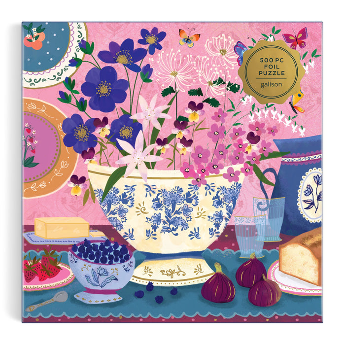 The Joy Laforme Art of the Table 500 Foil Piece Puzzle features a vibrant tableau with a blue and white bowl, flowers, fruit, cheese, figs, bread, and plates on a pink background—perfect for fans of Joy Laforme’s joyful art.
