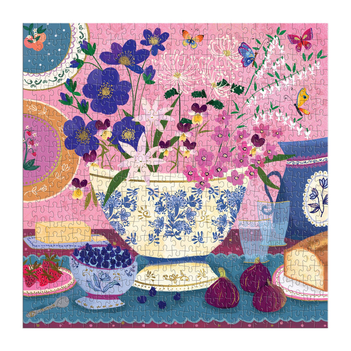 The Joy Laforme Art of the Table 500 Foil Piece Puzzle features a bright floral scene with a blue and white vase, plates, fruit, figs, bread on a blue tablecloth, plus butterflies and a bird against a pink backdrop.