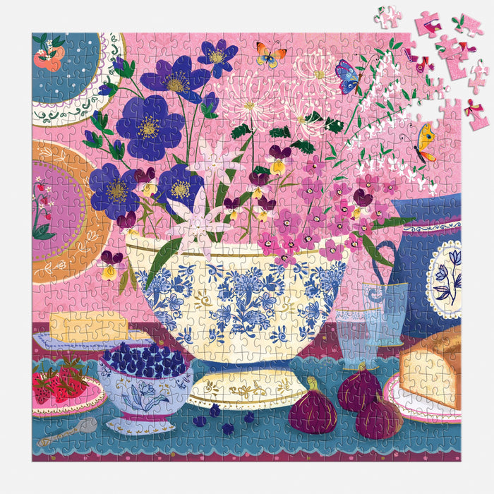 The Joy Laforme Art of the Table 500 Foil Piece Puzzle features a vibrant floral tableau with a blue and white vase, figs, berries, bread, and tableware—plus missing pieces in the upper right corner for extra intrigue.