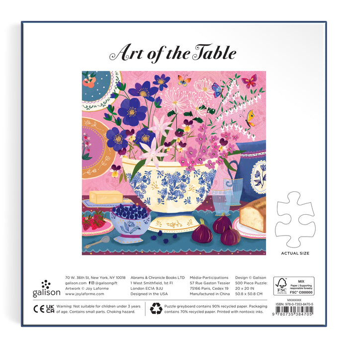 Inspired by Joy Laforme, the Art of the Table 500 Foil Piece Puzzle features a vibrant floral teacup with purple blooms, fruit, bread, cheese, and a blue teapot on a patterned table set against a pink background with butterflies and leaves.