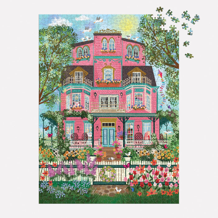 The Joy Laforme Capeside Victorian 1000 Piece Foil Puzzle depicts a grand Victorian mansion with balconies, trees, a white picket fence, and flowers. A few pieces remain unfinished in the upper right corner. Brand: Joy Laforme.