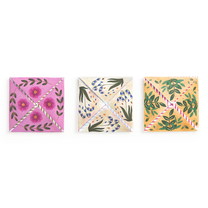 Three colorful origami fortune tellers, each with hand-painted pink, blue, and yellow floral designs, are arranged in a row—beautiful stationery inspired by Joy Laforme Charm Cards from Joy Laforme.
