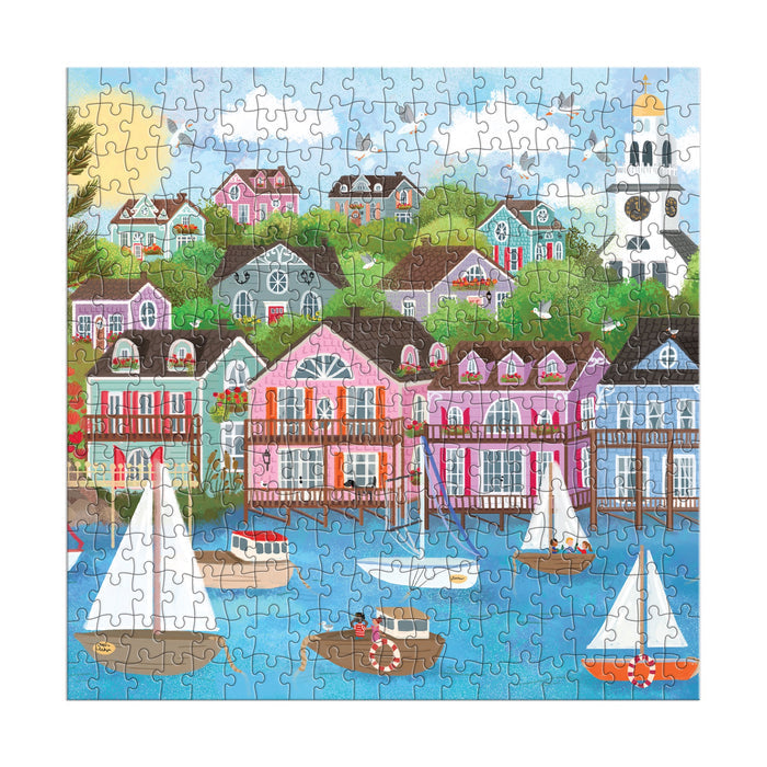 The Joy Laforme Harbor by the Sea 300 Piece Puzzle features a vibrant coastal village with colorful hillside houses, boats on blue water, lush trees, flowers, and a lighthouse under a bright sky. Brand: Joy Laforme.