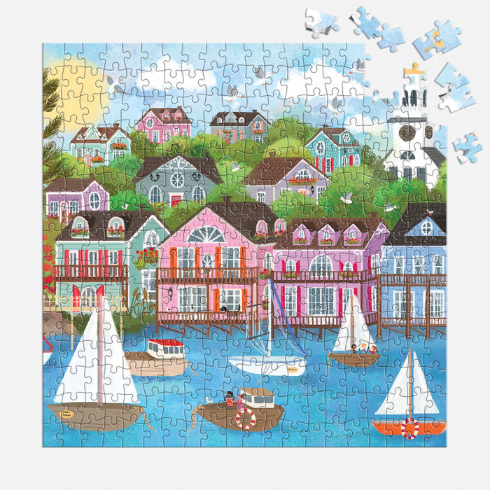The Joy Laforme Harbor by the Sea 300 Piece Puzzle shows sailboats on blue water with houses on a green hillside; several pieces are missing from the upper right in this cheerful puzzle by Joy Laforme.