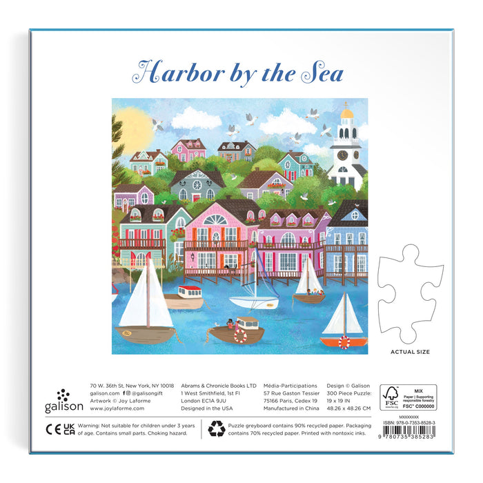 The Joy Laforme Harbor by the Sea 300 Piece Puzzle features a colorful harbor scene with sailboats, pastel hillside houses, trees, and a lighthouse. The puzzle box includes reference text and a sample piece for size. Brand: Joy Laforme.