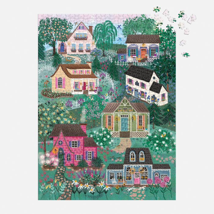 The Joy Laforme The Cottage Hills 1000 Piece Puzzle in a Square Box shows vibrant houses among gardens and trees, with a few missing pieces in the top right corner and loose pieces nearby on a white background.