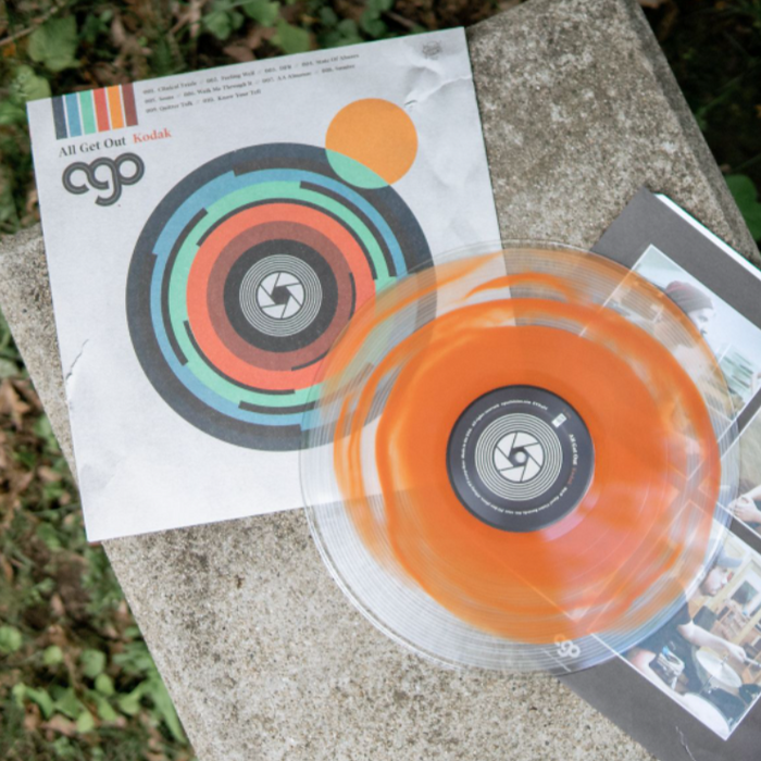 White vinyl jacket with artwork of a multicolored camera lens in the center. In the top left there is black lettering that says AGO. Above that is text that says ALL GET OUT, and next to it is orange lettering that says KODAK. On top of the jacket is a terracotta and clear colored vinyl. Both are on top of a granite surface.