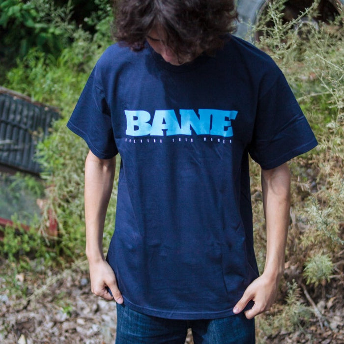 A person with medium-length brown hair looks down while wearing the iconic Bane T-Shirt, made from Gildan Hammer fabric, paired with jeans, and standing outdoors among greenery and dry plants.