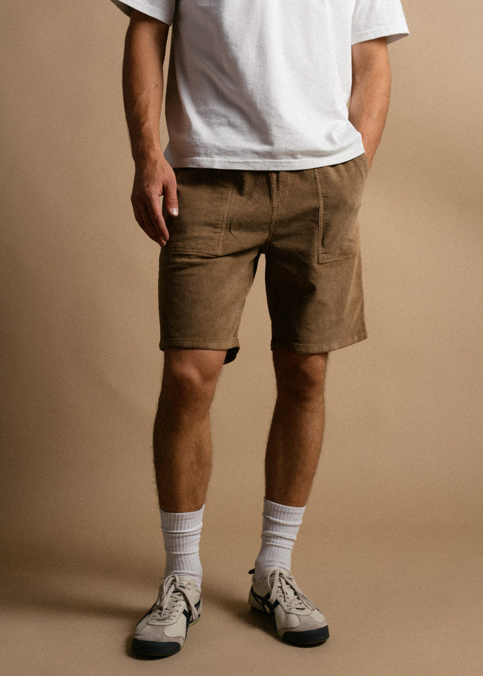 Man in white shirt, brown shorts, and sneakers stands against a beige background, hands in pockets, casual style.