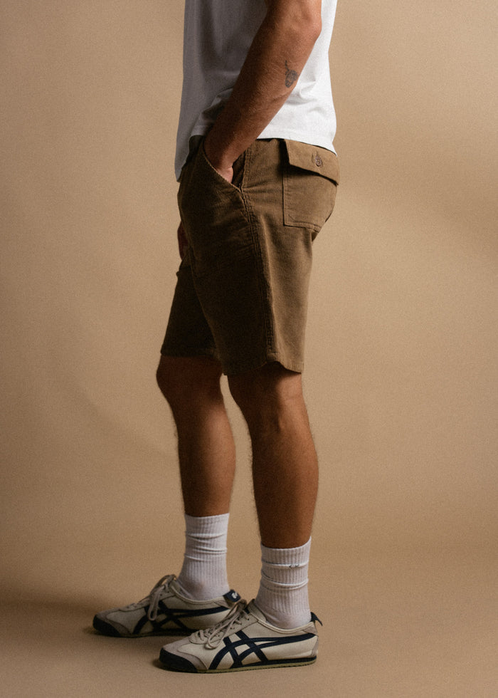Man in white shirt and brown shorts stands sideways, hands in pockets. Wearing white socks and sneakers against beige background.