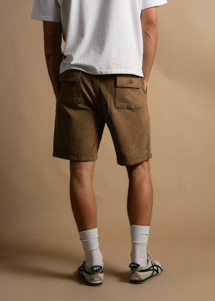 Man in white shirt, brown shorts, and sneakers stands on a beige background, facing away. Casual and relaxed style.