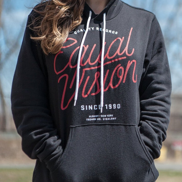 Black Equal Vision hoodie. Center of hoodie has "EQUAL VISION" printed in red cursive. Above has "QUALITY RECORDS" printed in white and below has "SINCE 1990" printed in white. Individual is modeling hoodie outside with blue sky and trees in the background.