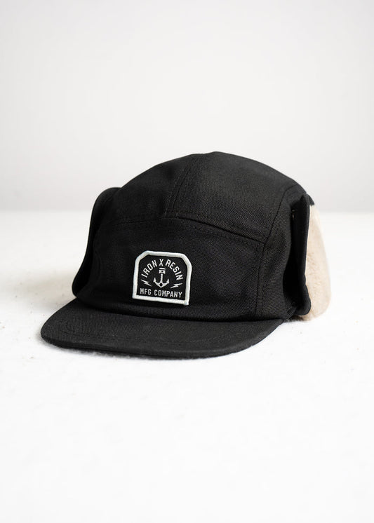 Black cap with ear flaps, featuring "Iron & Resin MFG Company" logo on front, displayed on a white surface.