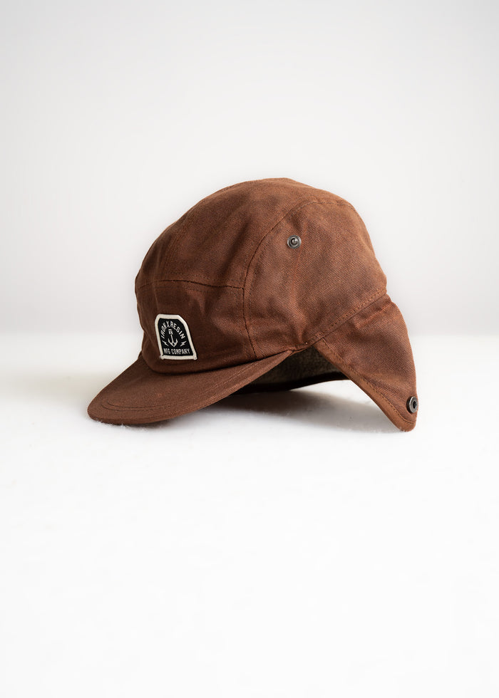 Brown waxed canvas cap with ear flaps and a patch logo, displayed on a white background, showcasing its rugged outdoor style.