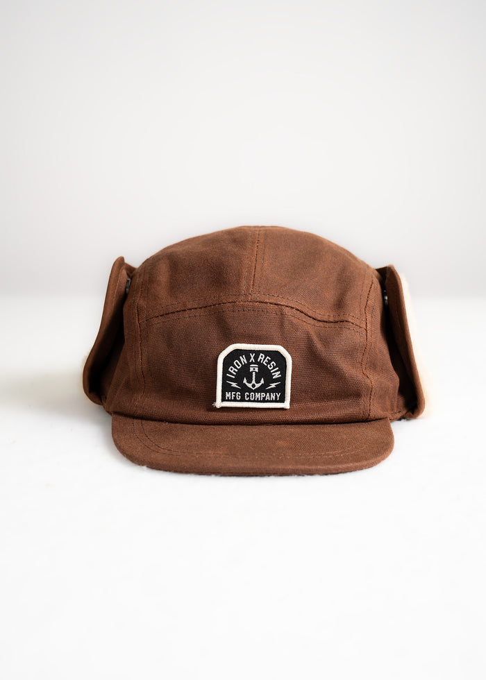 Brown cap with ear flaps, featuring an Iron & Resin MFG Company patch on the front, set against a plain white background.