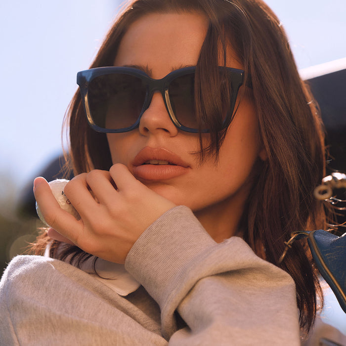 lifestyle - female holding a golf ball outdoors wearing diff eyewear featuring the meredith square oversized sunglasses with a clubhouse blue acetate frame and grey gradient polarized lenses front view