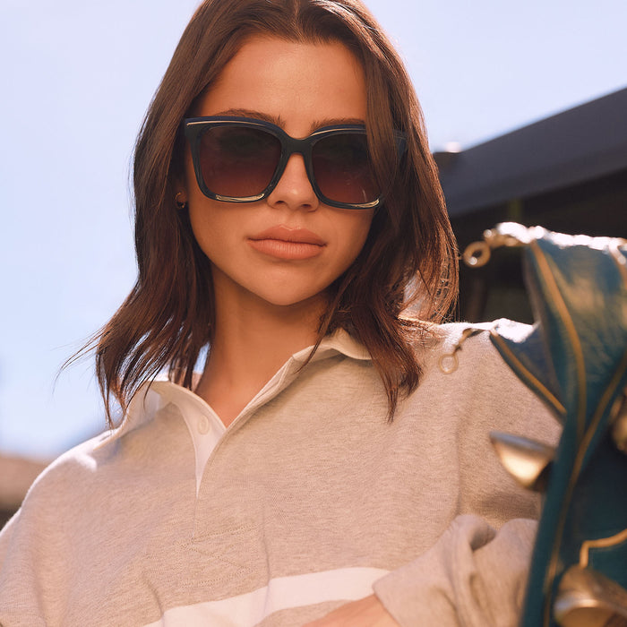lifestyle - female holding a golf bag outdoors wearing diff eyewear featuring the meredith square oversized sunglasses with a clubhouse blue acetate frame and grey gradient polarized lenses front view