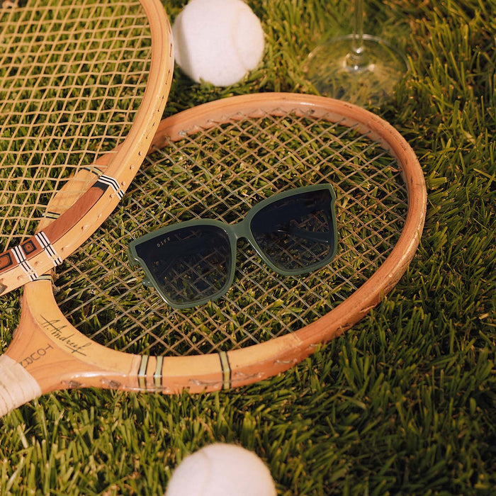 still life - diff eyewear meredith ii square oversized sunglasses with a fairway green acetate frame and grey gradient lenses resting with tennis rackets on the lawn