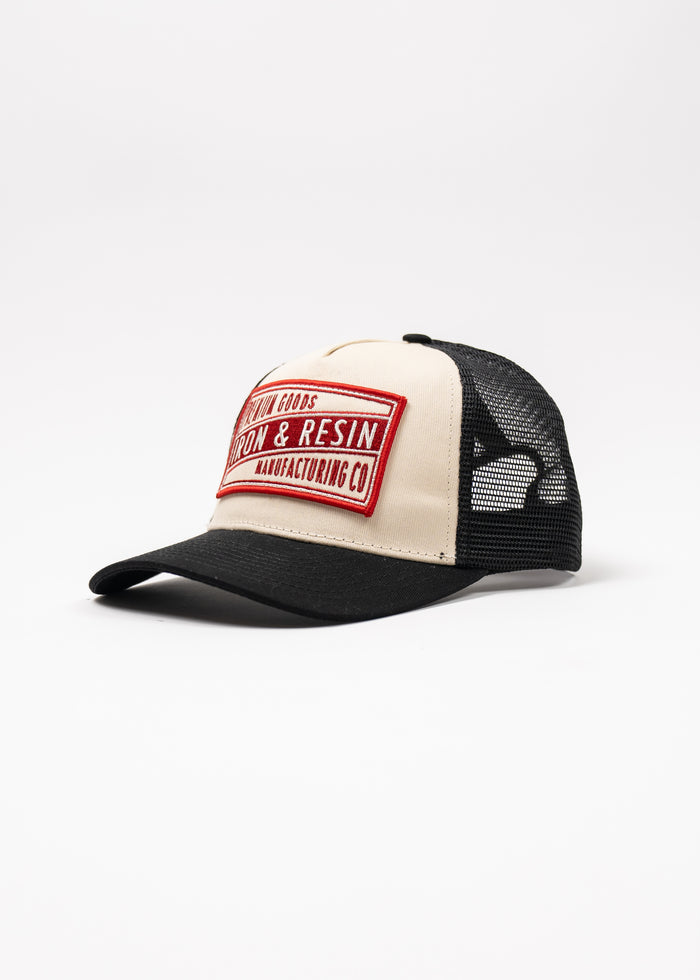 Trucker cap with black mesh, beige front, and red "Iron & Resin Manufacturing Co" patch, set against a white background.