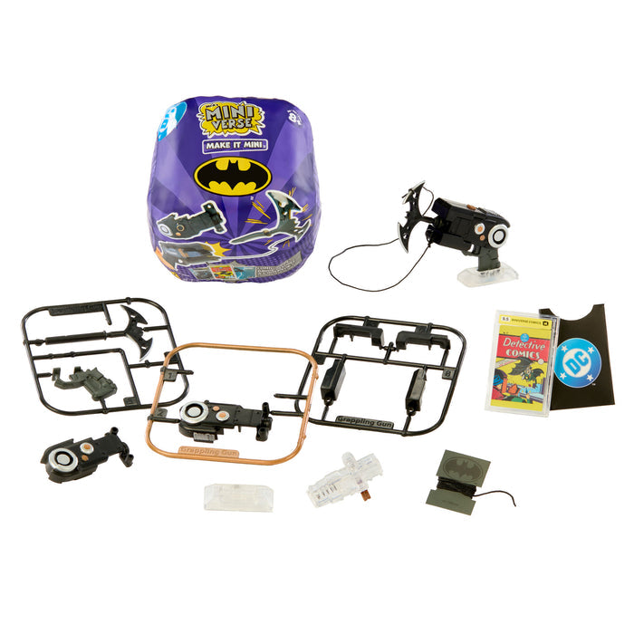 A Batman-themed toy set laid out on a white background, including a purple packaged round item, a small black flying 'bat' with a wire, several black plastic parts on frames, a small book,  and a few small accessories.