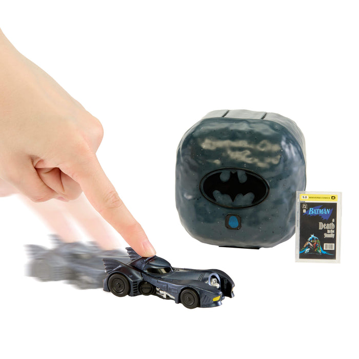 A Batman toy car with motion blur from being touched, shown next to a dark gray Batman-themed cube control and a Batman Death Knight book, on a white background.