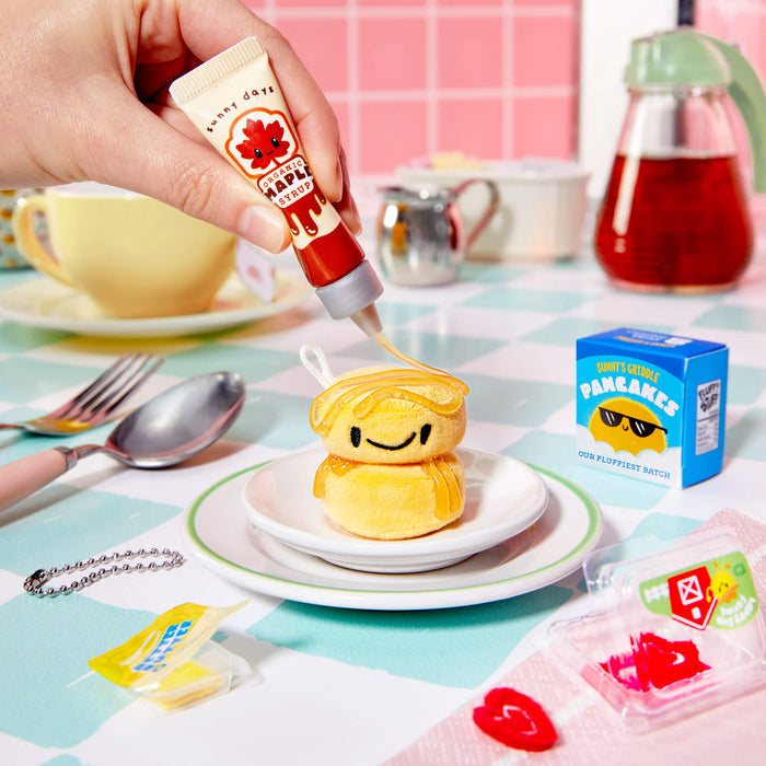 Hand squeezing a tube of "syrup" onto pancakes on a colorful tablecloth with various items around.
