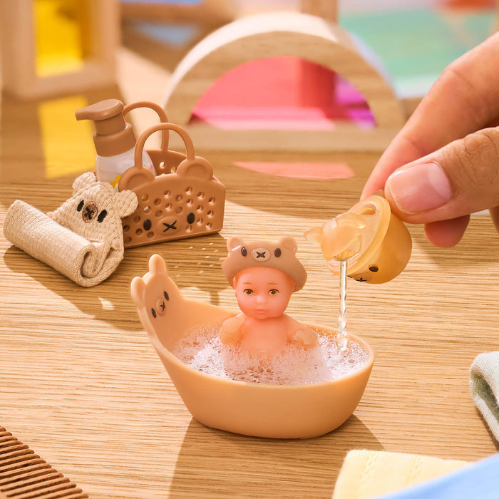 Mini toy baby in a bathtub with a hand pouring water, surrounded by bath accessories on a wooden surface.