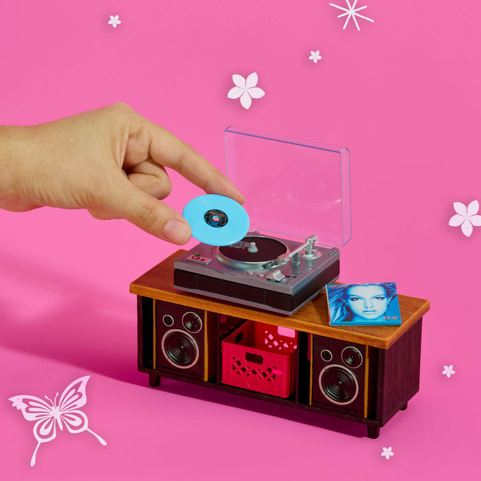 Miniature record player with a hand placing a vinyl record on it against a pink background with decorative elements.