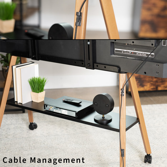 A close-up of a black cable management clip mounted on a wooden rolling TV stand in a living room setting. The lower shelf of the stand holds a cable modem, a smart speaker, and a small potted plant, with a TV mounted above.
