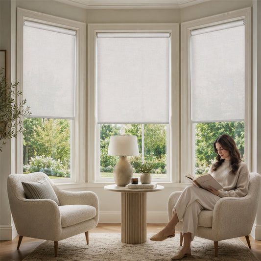 A woman sits in a cream upholstered armchair in a bright corner room, reading a book beside a small round table with a lamp and potted plants. Two more matching armchairs are visible, and the windows above have light-colored roller shades partially drawn.