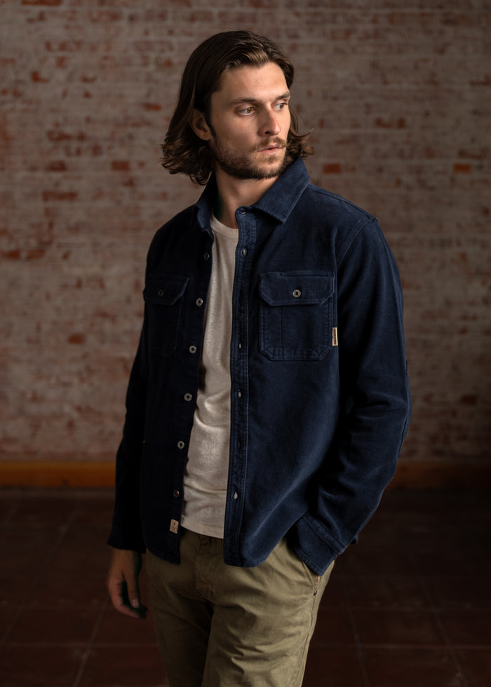 Man in a navy jacket and white shirt stands against a brick wall, looking to the side with a thoughtful expression.