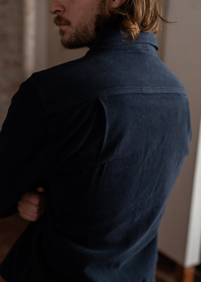 Man with long hair in a navy shirt, seen from behind, standing indoors with arms crossed, looking to the side.