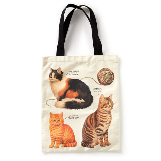 The Brass Monkey Moody Cats Canvas Tote is a cream-colored bag with black handles, featuring whimsical illustrations of a calico, orange tabby, and brown tabby playing with yarn—each labeled for extra charm.