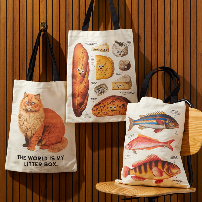Three illustrated Brass Monkey totes hang on a wooden wall: the Moody Cats Canvas Tote with The world is my litter box, one with cheese and bread labels, and another with three colorful fish. The fish tote rests on a chair.