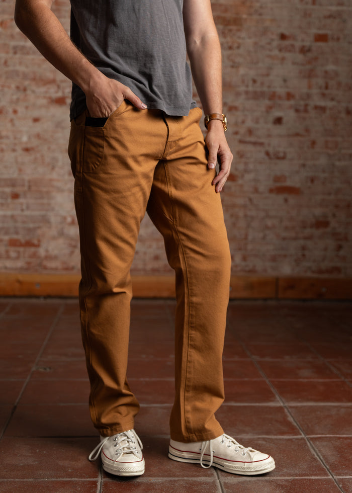 Person in brown pants and white sneakers stands on a tiled floor, hand in pocket, against a brick wall. Casual style.