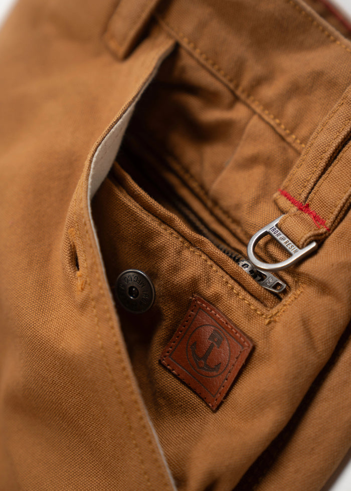 Close-up of brown pants pocket with a metal zipper, button, and leather anchor patch, highlighting rugged, detailed craftsmanship.
