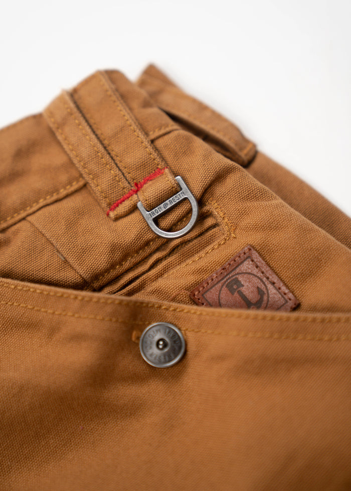 Close-up of brown pants with a metal button, red stitching, and a branded D-ring detail on the belt loop.