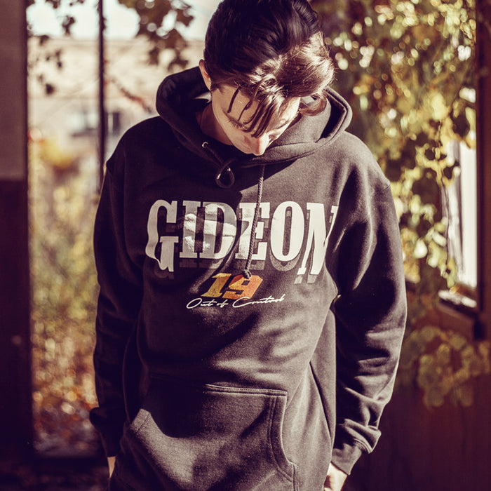 Black pullover hoodie with GIDEON written across the chest in white font. Below that the number 19 is written in a yellow and orange gradient font. Below that there is small white text that says OUT OF CONTROL. An individual is modeling the hoodie and standing in a forest with trees.