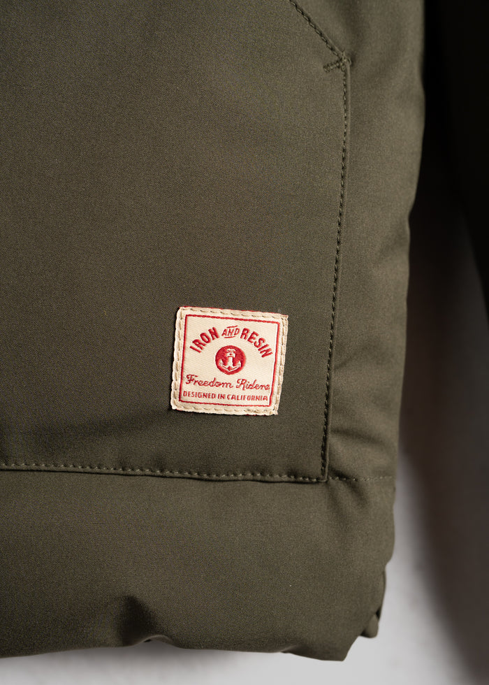 Close-up of an olive green jacket with a red and white "Iron and Resin" label, reading "Freedom Riders, Designed in California."